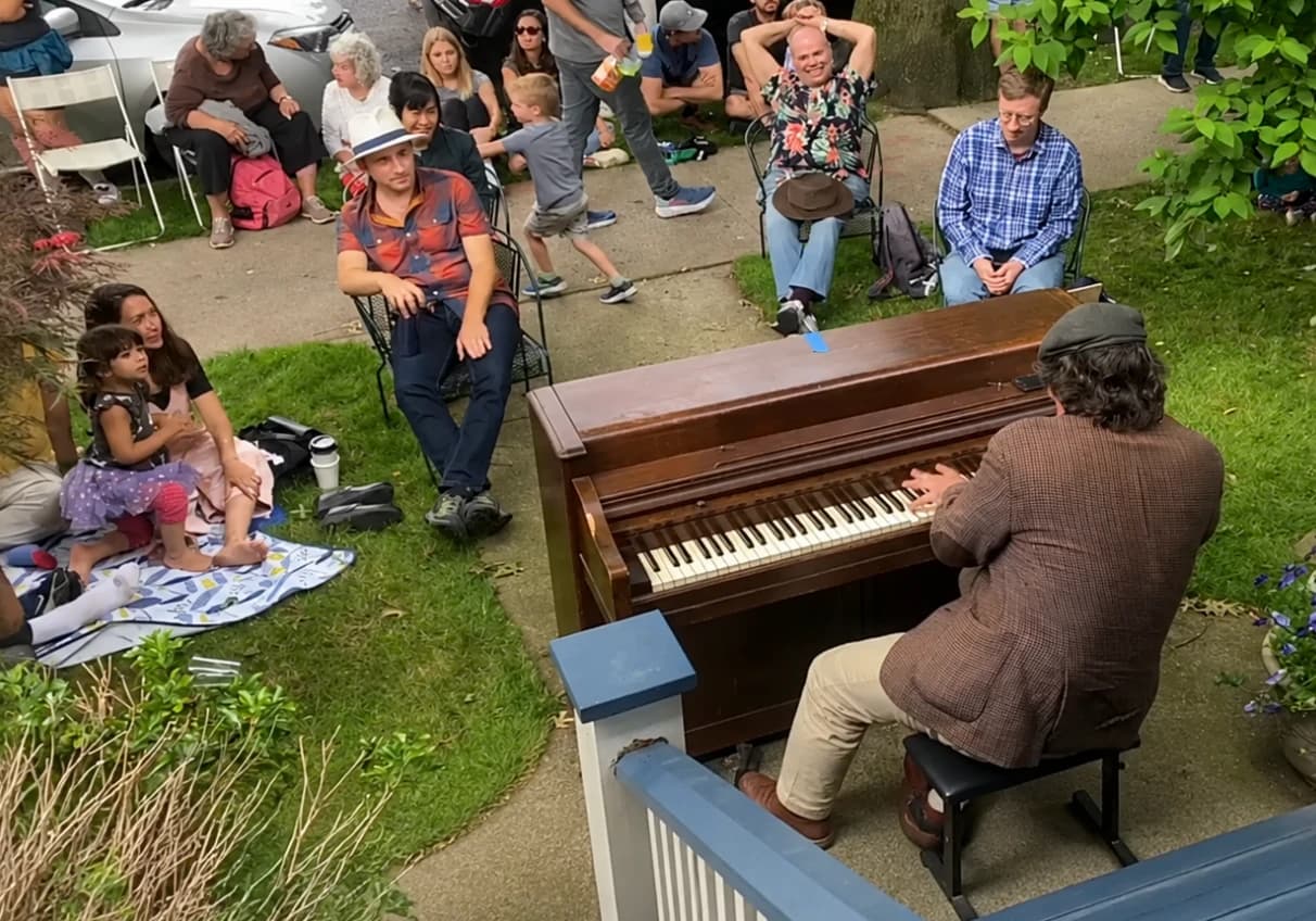 A pianist playing an upright piano on a Ditmas Park front lawn, with neighbours listening from folding chairs, a picnic blanket and the sidewalk